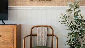 A room with a wooden chair with a patterned cushion, a potted plant, a wall-mounted light fixture, and a framed painting above a white paneled wall.