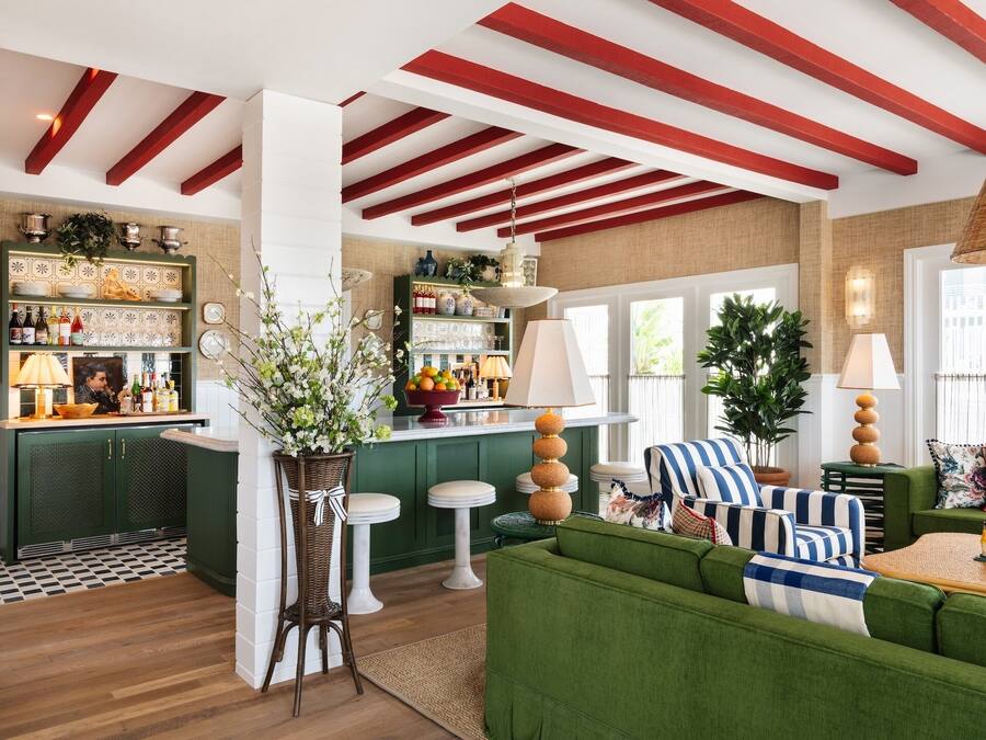 Zona de bar con armarios verdes, taburetes blancos y una barra. Hay un sofá verde con cojines a rayas y una mesita con una lámpara.