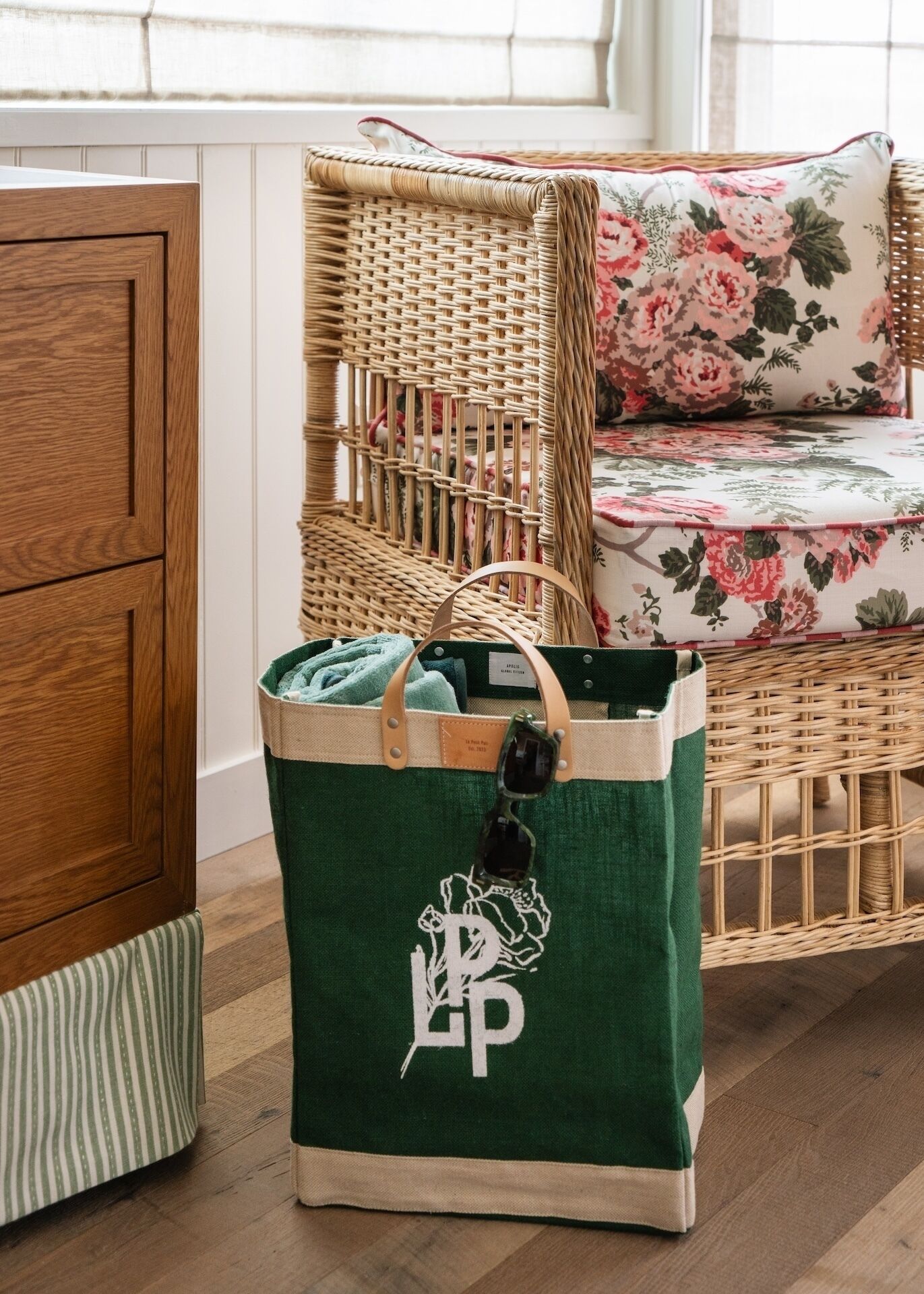 Ein Korbstuhl mit floralen Kissen, eine grüne Tragetasche mit Logo und ein gestreiftes Kissen auf Holzdielen.