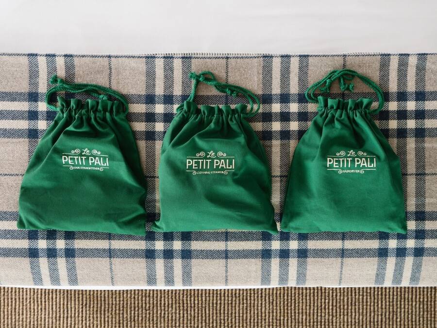 Tre sacchetti verdi con coulisse e marchio 'Petit Pali' su sfondo di tessuto a quadri.