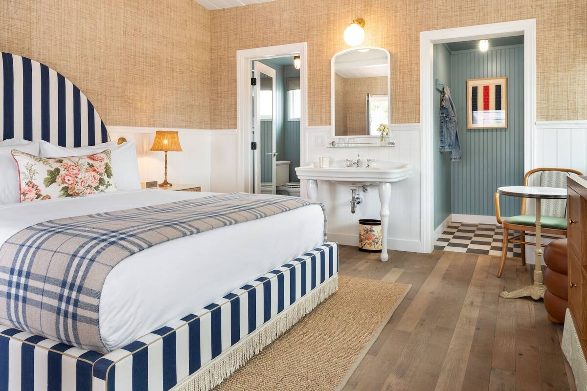 A bedroom with a large bed, a bedside table, a lamp, a bathroom with a sink and mirror, and a chair.