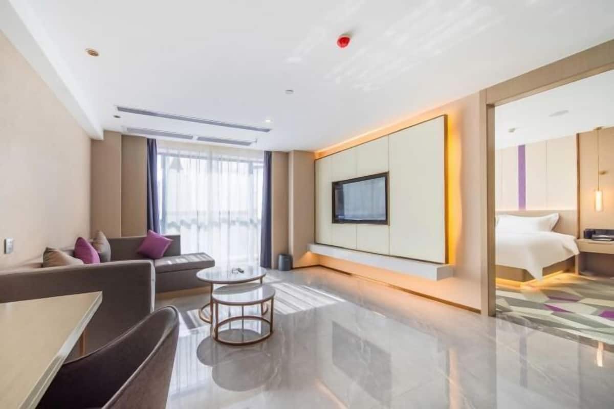 A modern hotel room with a sofa, a small round table, a TV mounted on the wall, a bed, and a large window with curtains.