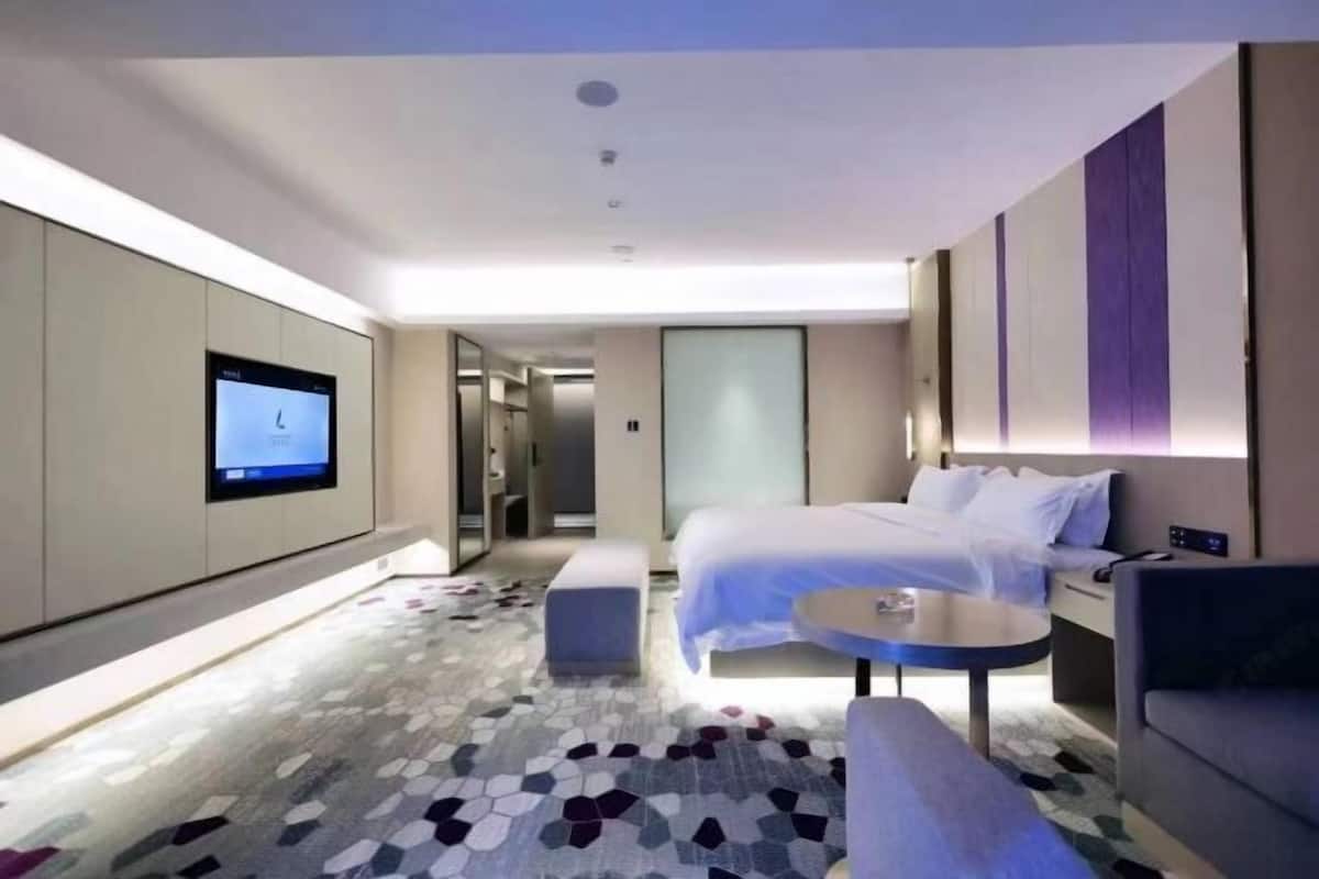 A modern hotel room with a bed, a small table, a sofa, a TV mounted on the wall, and a patterned carpet.