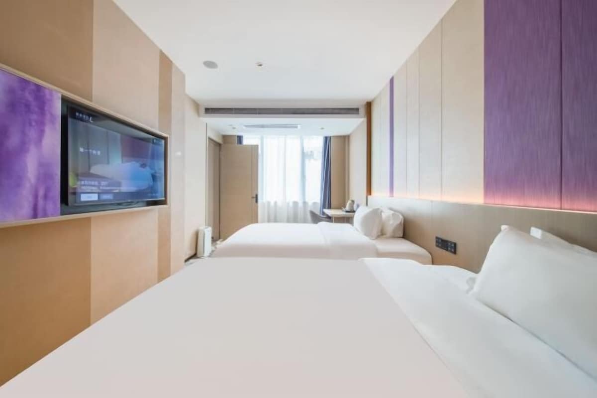 A modern hotel room with a large bed, a flat-screen TV mounted on the wall, and a view of the city through the window.