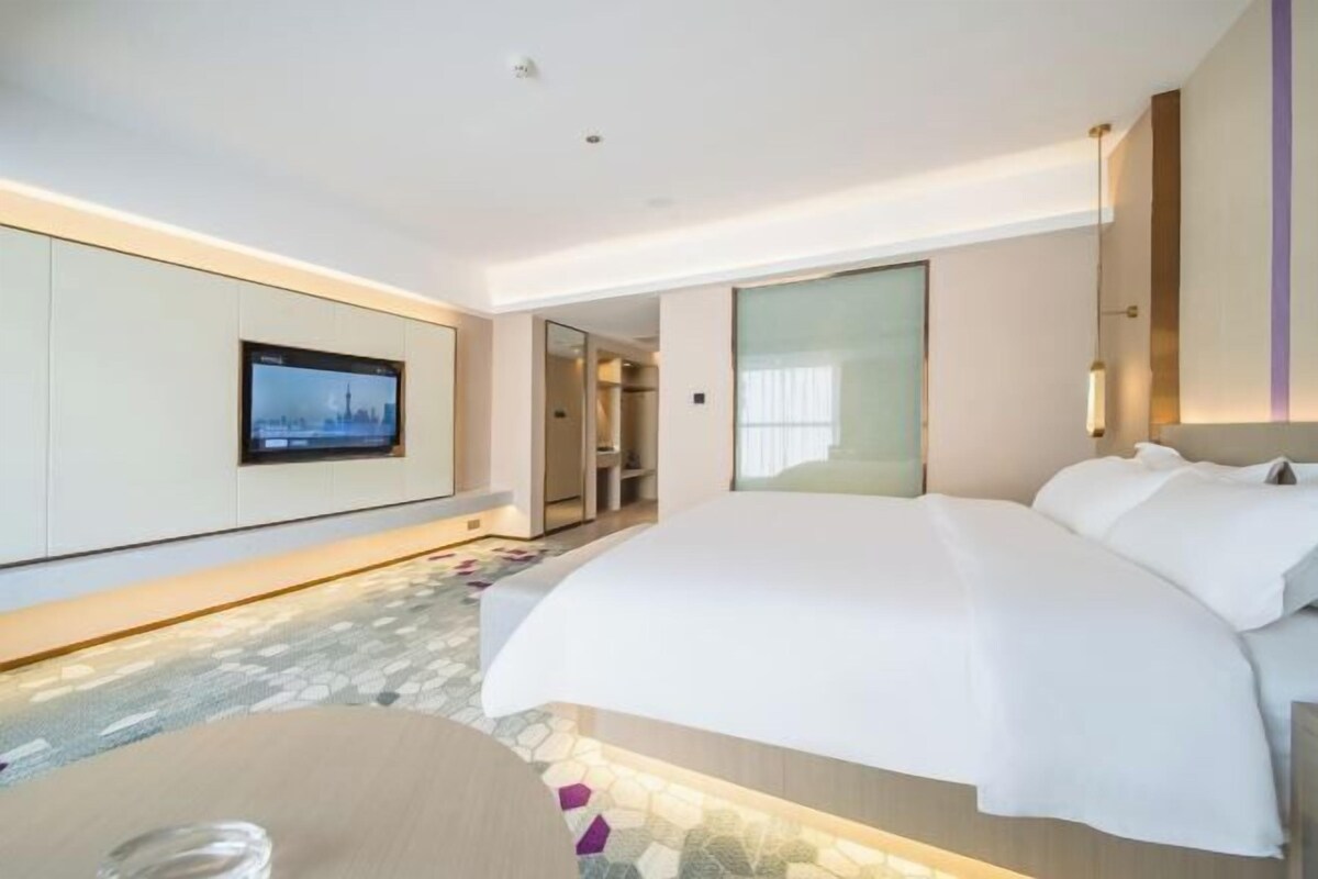 A modern hotel room with a large bed, a flat-screen TV mounted on the wall, a glass door leading to a bathroom, and a stone-patterned floor.