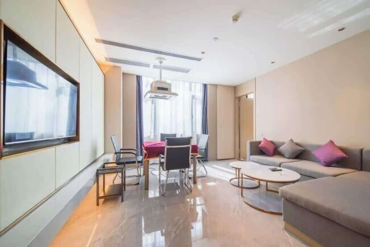 A modern hotel room with a sofa, a dining table with chairs, a coffee table, and a large window.