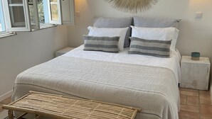 A neatly arranged bedroom with a bed, bedside tables, a wall-mounted lamp, and a window with open shutters. - Jasmin House | Herdade d' Amoreira (Aljezur)