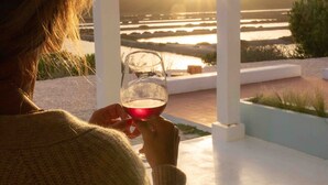 A person holding a glass of red wine at a table with a view of the sunset over a body of water. - Jasmin House | Herdade d' Amoreira (Aljezur)