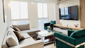 Una sala moderna con un sofá gris, un sillón verde azulado, una mesa de centro de vidrio y un televisor montado en la pared.