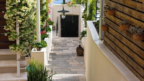 Un étroit sentier de pierre mène à une porte noire, flanquée de murs blancs et de balustrades en bois ornées de pots de plantes.