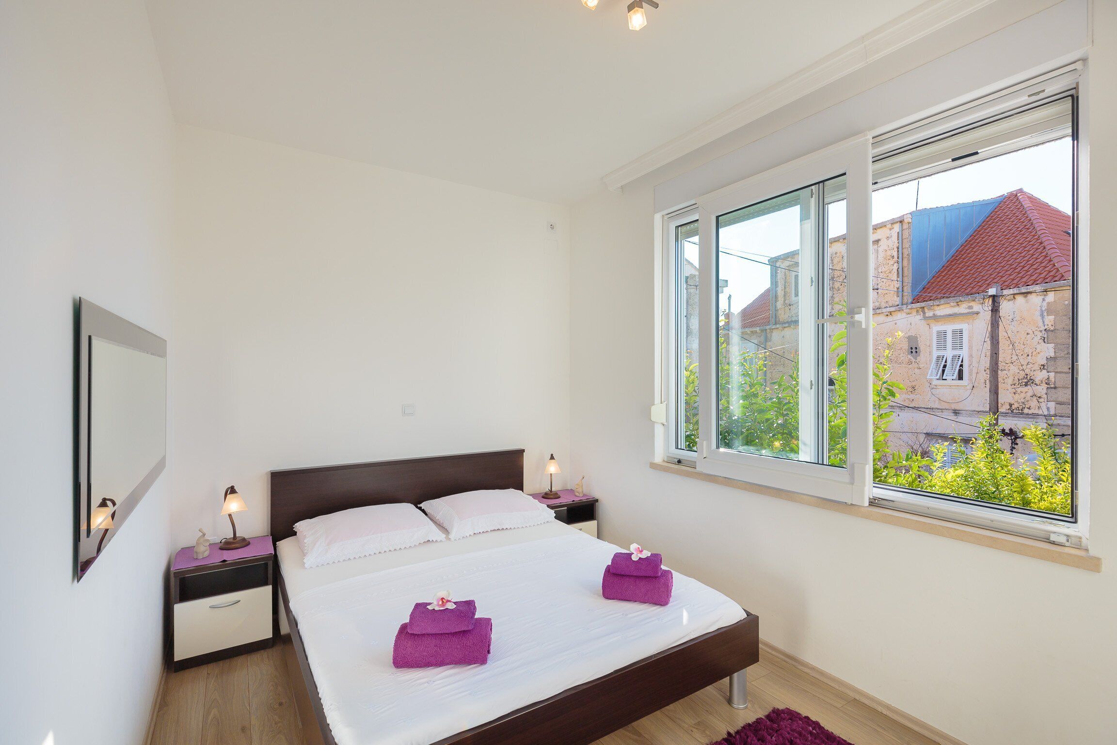 A bedroom with a bed, two bedside tables, a window, and a view of buildings outside.