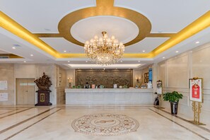 A hotel lobby with a reception desk, a chandelier, a decorative wall, and a patterned floor.