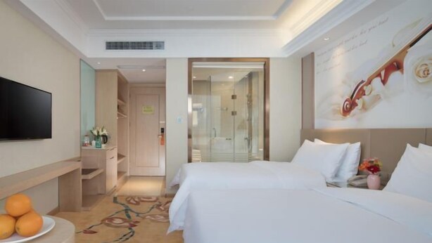 A hotel room with a bed, a TV, a small table with fruit, and a bathroom visible through a glass door.