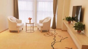 A hotel room with two white armchairs, a small round table with a vase of flowers, a large window with sheer curtains, a potted plant on a shelf, and a floral carpet design. - Vienna Hotel (Feidong Dongcheng High-speed Railway Station) (Feidong)