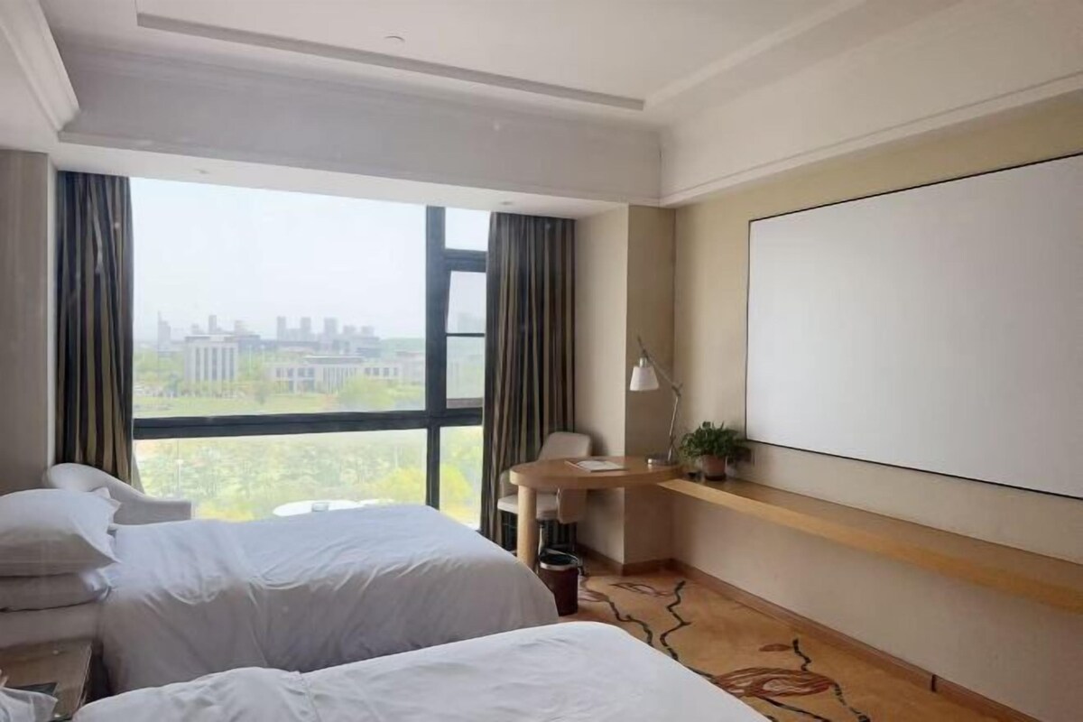 Habitación de hotel con dos camas, un escritorio, una silla, un ventanal amplio con vistas a la ciudad y una pantalla montada.
