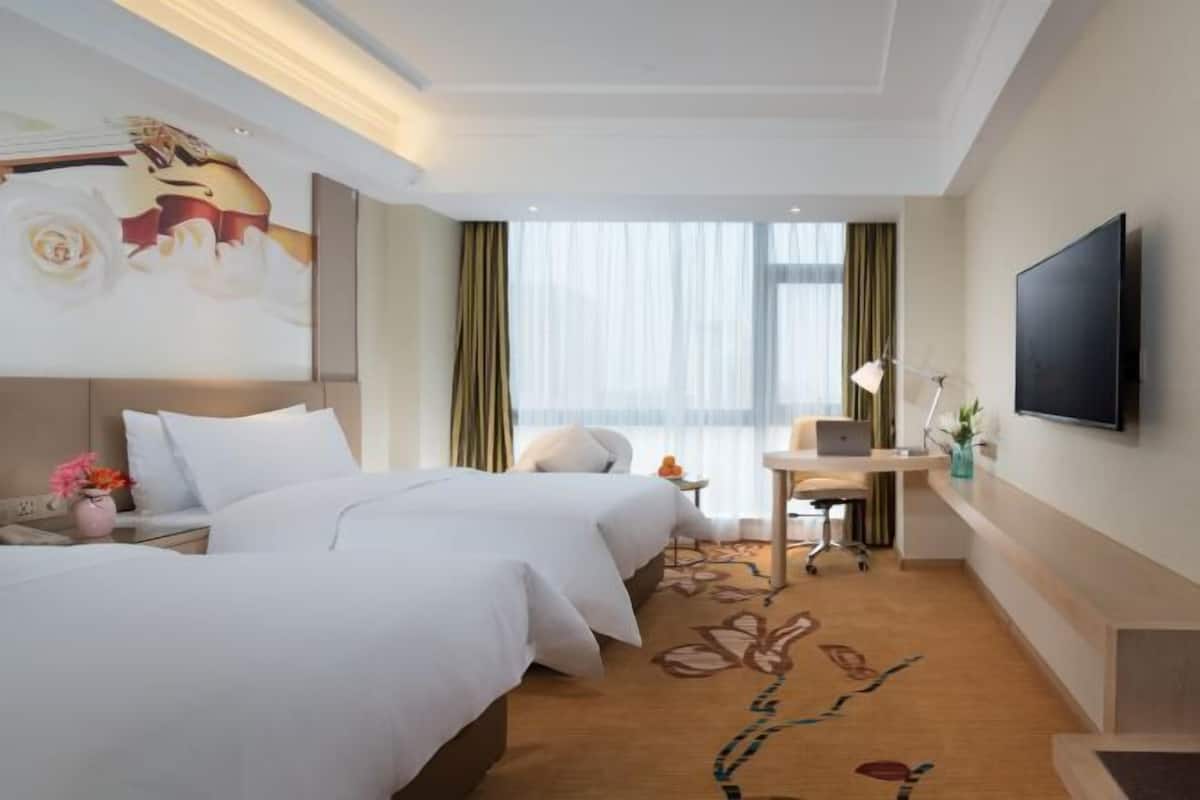 A hotel room with two beds, a desk with a laptop, a flat-screen TV, and a large window with curtains.