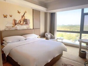 A hotel room with a large bed, a chair, a small table, and a view of the city through large windows. - Vienna Hotel (Feidong)