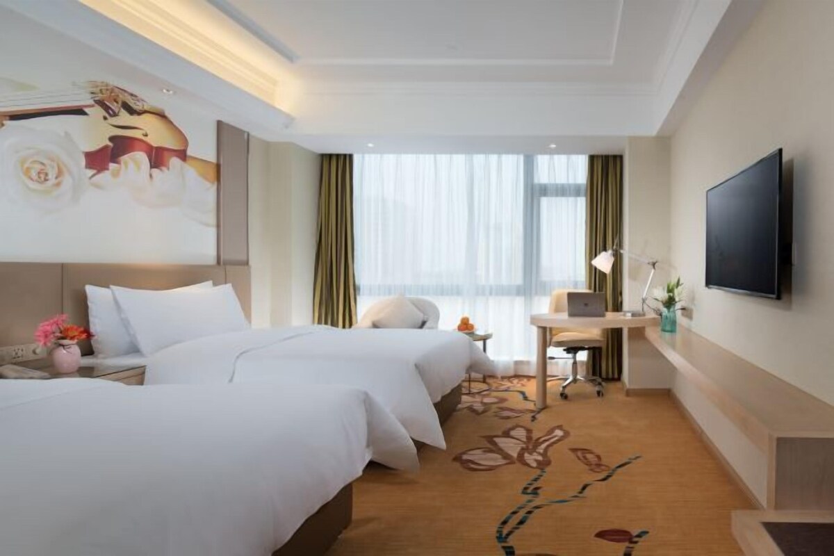 A hotel room with two beds, a desk with a laptop, a flat-screen TV, and a large window with curtains.