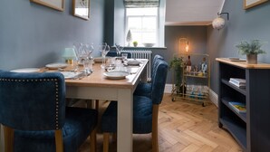 A dining area with a wooden table set for six, flanked by blue upholstered chairs. A globe lamp hangs above the table, and a plant is on a shelf to the right. - Wansfell at Lipwood House (Windermere)