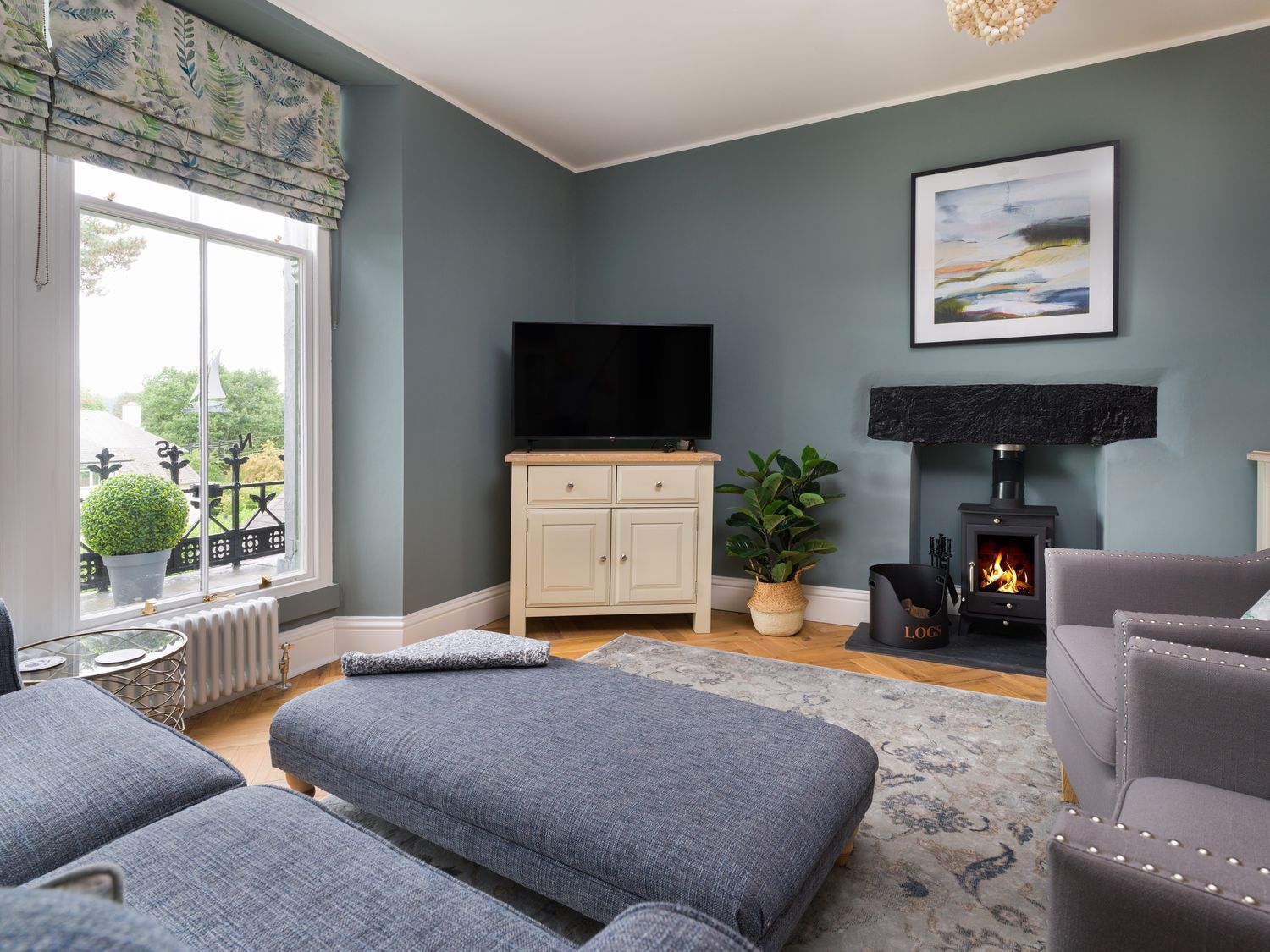 A living room with a grey sofa, a grey armchair, a TV on a wooden cabinet, a fireplace, and a large window with a view of greenery.