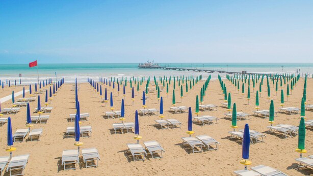 Une plage bordée de rangées de transats et de parasols, une jetée en arrière-plan et un drapeau rouge sur la gauche.