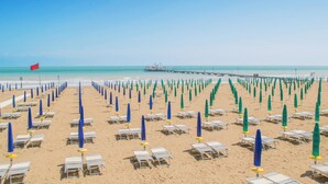 A beach with rows of sun loungers and umbrellas, a pier in the background, and a red flag on the left.