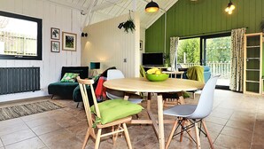 En rummelig stue med højt til loftet, et spisebord med stole, et fjernsyn og udsigt til grønne omgivelser.