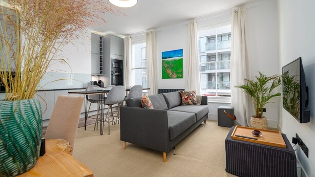 Un apartamento moderno con sala de estar, comedor y kitchenette. Incluye un sofá gris, una mesa de comedor con sillas, un televisor montado en la pared y ventanas grandes con cortinas.