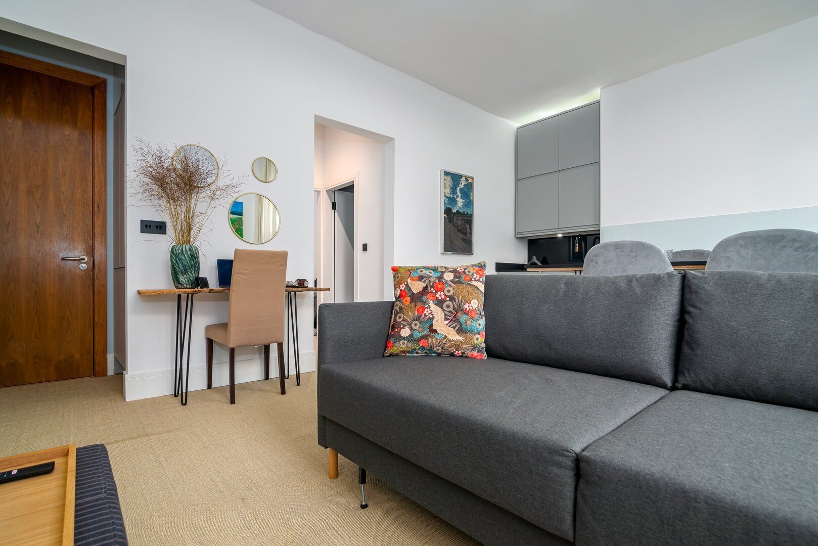 A modern living room with a grey sofa, a wooden door, a desk with a chair, and a small table with a vase.