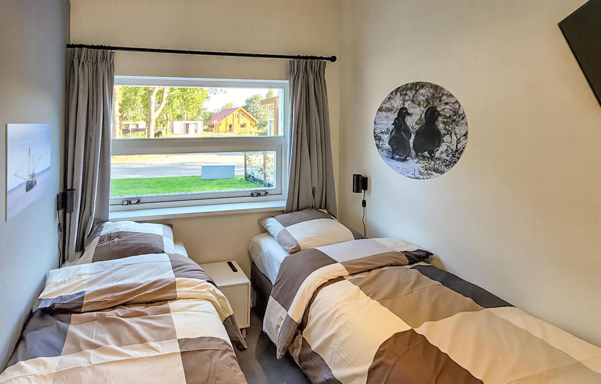 A bedroom with two beds, a window with curtains, a wall-mounted lamp, and a picture of birds.