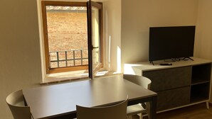 A small, well-lit room with a dining table and chairs, a television on a stand, and a window with a view of a brick wall. - San Donato residence (Montepulciano)