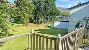 A wooden deck with a railing, a metal bench, and a view of a grassy yard with trees and a building in the background. - The Prince - UK42204 (Glendaruel)