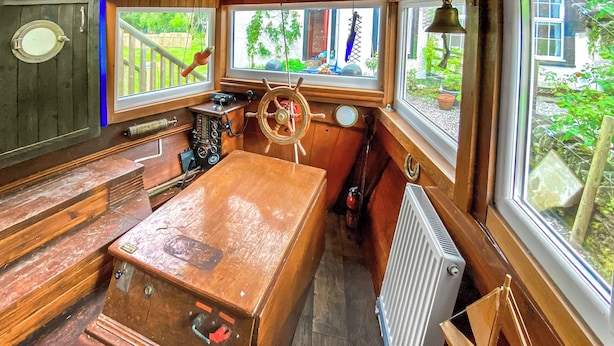 A cabin interior with a wooden table, a steering wheel, a radio, and a window with a view of greenery. - The Prince - UK42204 (Glendaruel)