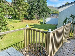 A wooden deck with a railing, a metal bench, and a view of a grassy yard with trees and a building in the background.