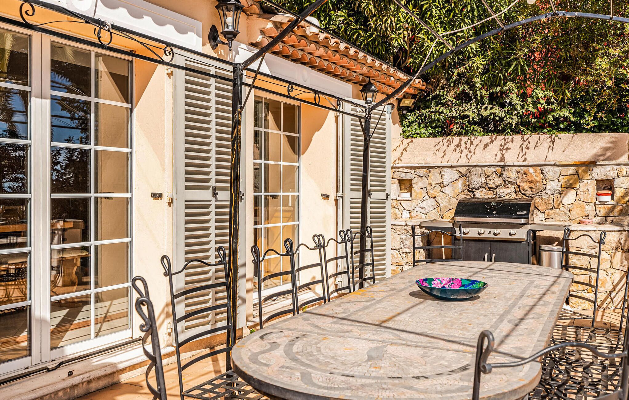 Une terrasse extérieure avec une table en pierre, des chaises en métal, un barbecue et une vue sur un mur de pierre et de la verdure.