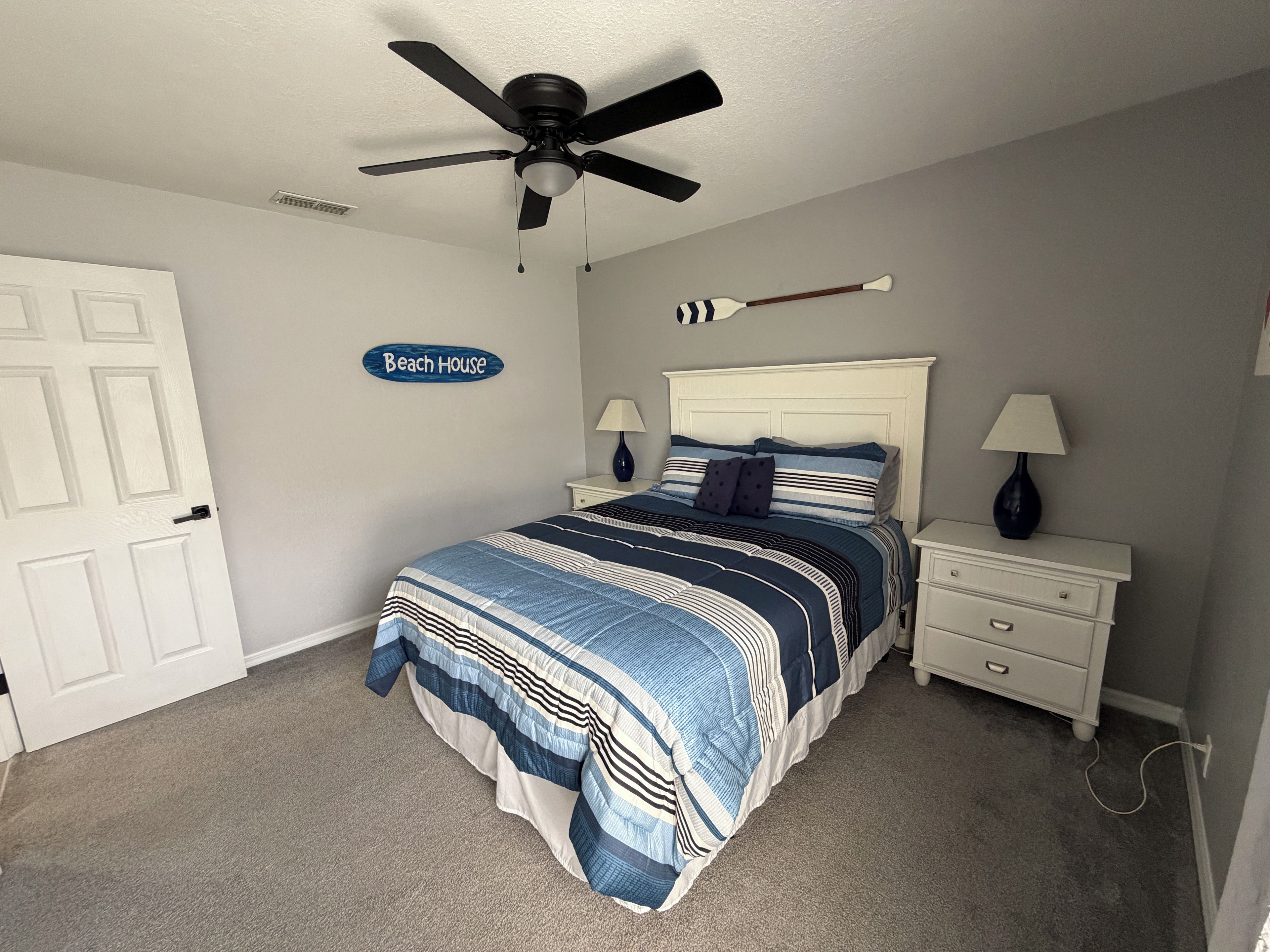 A bedroom with a bed, a ceiling fan, a nightstand with lamps, a wall sign, and a door.