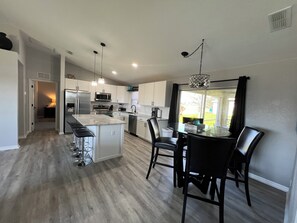 A modern kitchen with a central island, bar stools, and a dining area with chairs and a table. - Beachy Modern Vacation Home (Cape Coral)