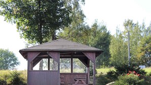 Un kiosque en bois avec une table de pique-nique, entouré de verdure et d’arbres.