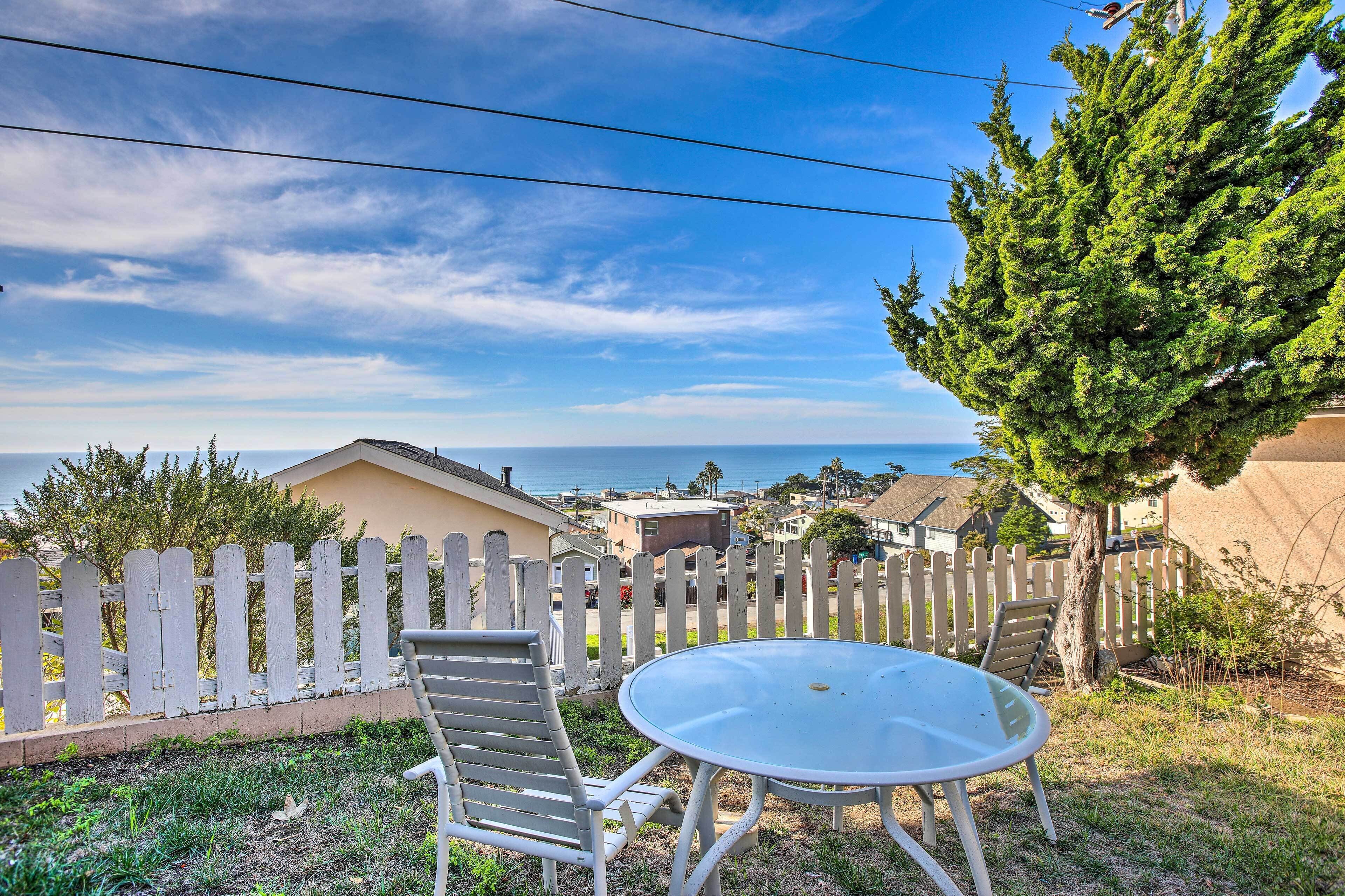 A patio with a round table and two chairs, a white picket fence, and a view of the ocean.