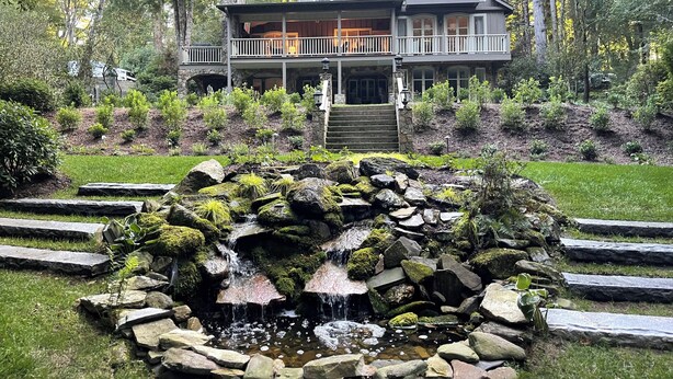 A two-story house with a stone staircase leading to a balcony, surrounded by lush greenery and a small waterfall feature.