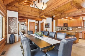 A spacious dining area with a long table and multiple chairs, a kitchen with wooden cabinets, and a ceiling with exposed beams.