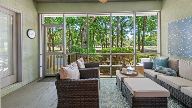 A sunlit patio with wicker furniture, a clock, and a view of a wooded area through large windows. - 3BR/3BA Ground Level ,Gated Pawleys Plantation (Pawleys Island)