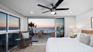 Un dormitorio con una cama grande, ventilador de techo, vista al océano y balcón con asientos.