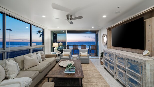 Un salón espacioso con un amplio ventanal con vista al océano, un ventilador de techo y un mueble de madera con un televisor de pantalla plana.