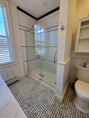 A bathroom with a glass-enclosed shower, white tiled walls, a patterned floor, and a toilet with a shelf above it.