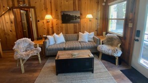 Een gezellige woonkamer met houten wanden, een gestreepte bank, twee fauteuils, een salontafel en een boslandschap op een schilderij.
