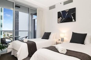 Two single beds with white linens and dark brown headboards, a bedside table, and a large window offering a city view.