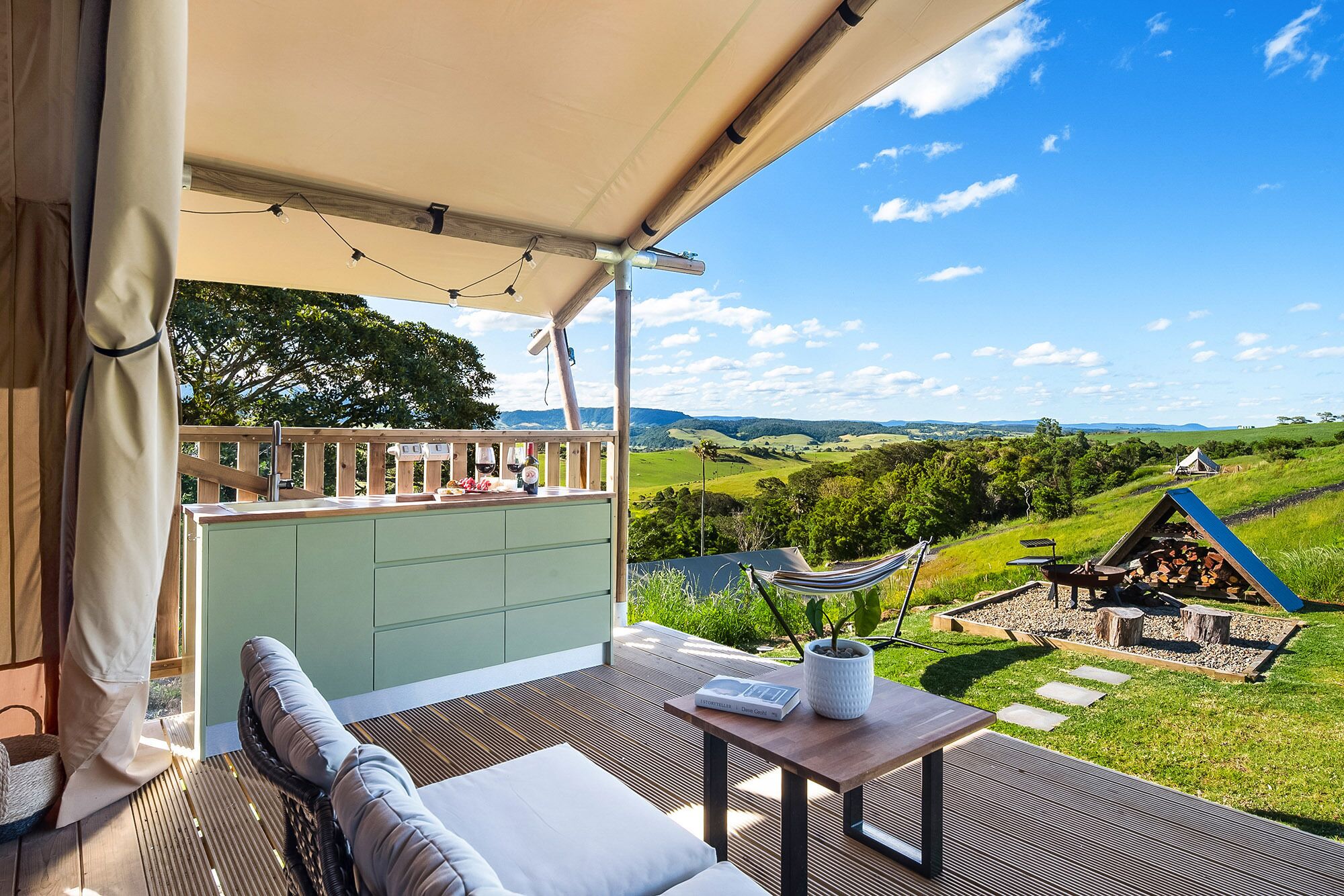 A balcony with a view of rolling hills, a hammock, a table with a book, and a small barbecue area.
