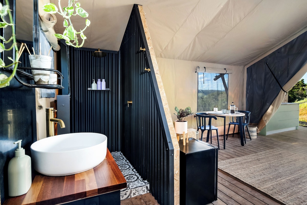 A modern bathroom with a large sink, a shower, and a wooden vanity. There is a small table with chairs outside the tent.