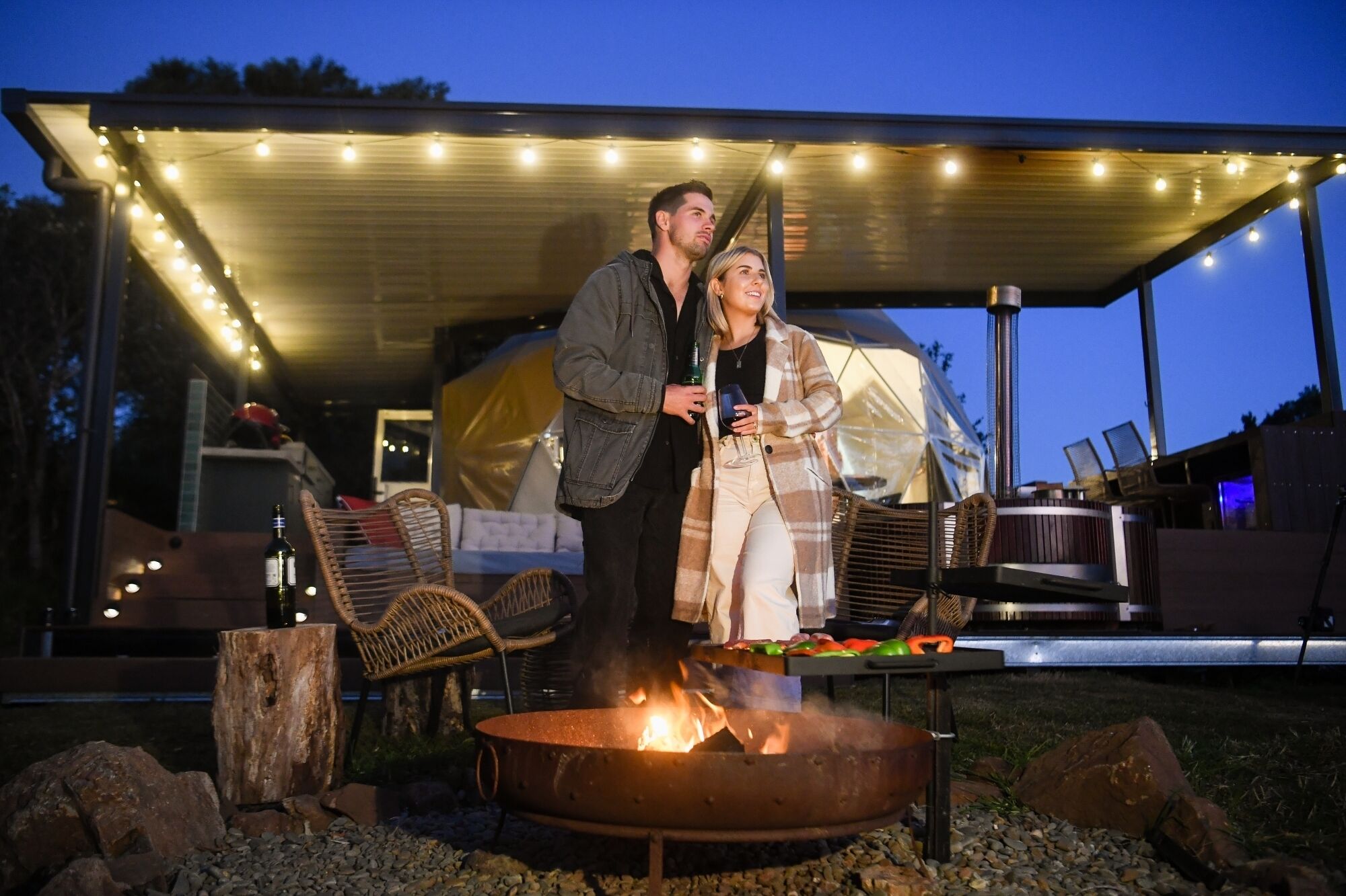 Un couple se tient près d’un foyer extérieur, avec un espace salon couvert et des guirlandes lumineuses en arrière-plan.